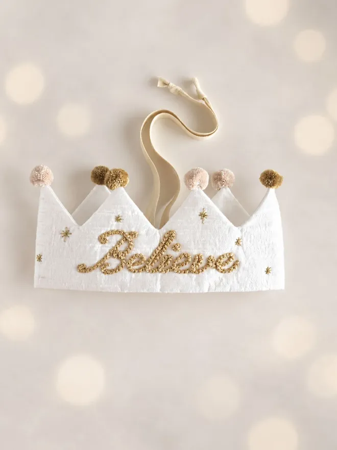 Handmade white silk Believe Christmas crown with metallic gold embroidery, embroidered stars, soft pom-poms and an ivory ribbon tie.