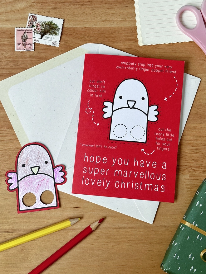A colour in Christmas card with a robin finger puppet design lays on a white envelope on a wooden desk.