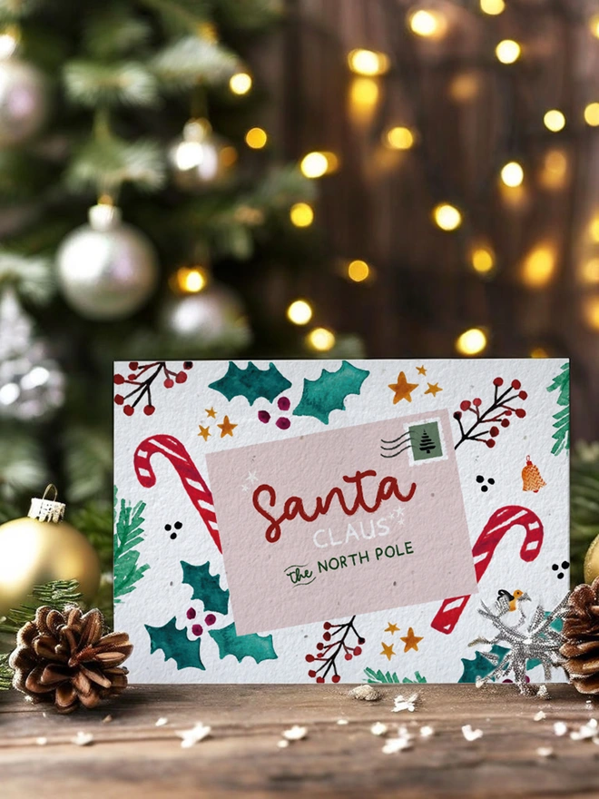 In the centre of the image is a card with a postcard addressed to Santa Clause surrounded by holly, star and candy cane illustrations. The card is set within a Christmas scene