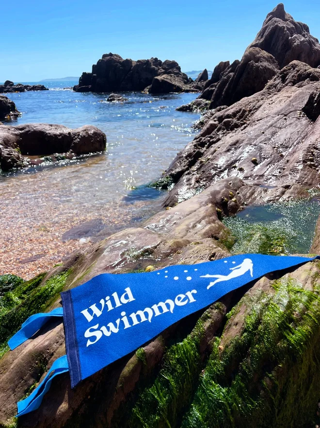 A turquoise sea leads in to a cove of rocks, the sun shining on them, on the rock is laid a bright royal blue felt pennant flag, in white text is written Wild Swimmer with a silhouette of a lady swimming next to it