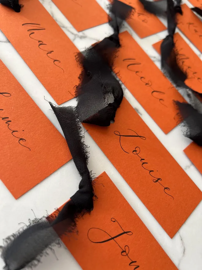Personalised Calligraphy Rust Orange Place Card, orange place cards with black chiffon ribbons grouped together on a white surface. 