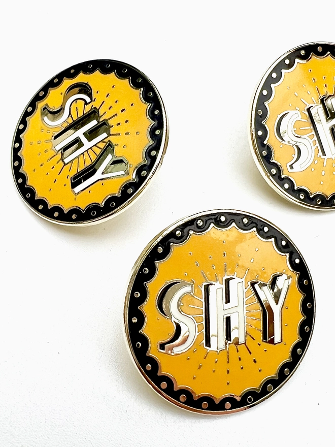 A close up of three SHY cloisonne brooches, they are mustard, black, white and brass coloured. 