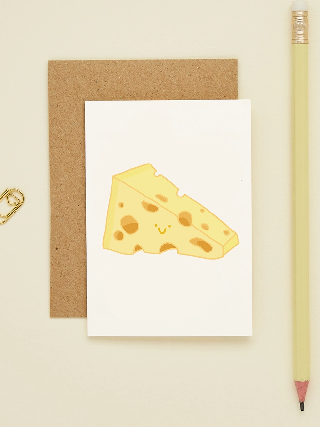 Mini greeting card featuring a cute wedge of cartoon cheese with a smiling face, styled with a kraft envelope and yellow pencil on a cream background.