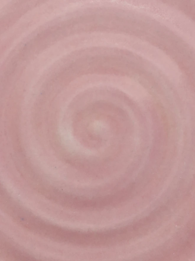 A close-up of a handmade ceramic glaze swatch in soft blush pink, showing a smooth concentric swirl texture with a delicate, even tone throughout