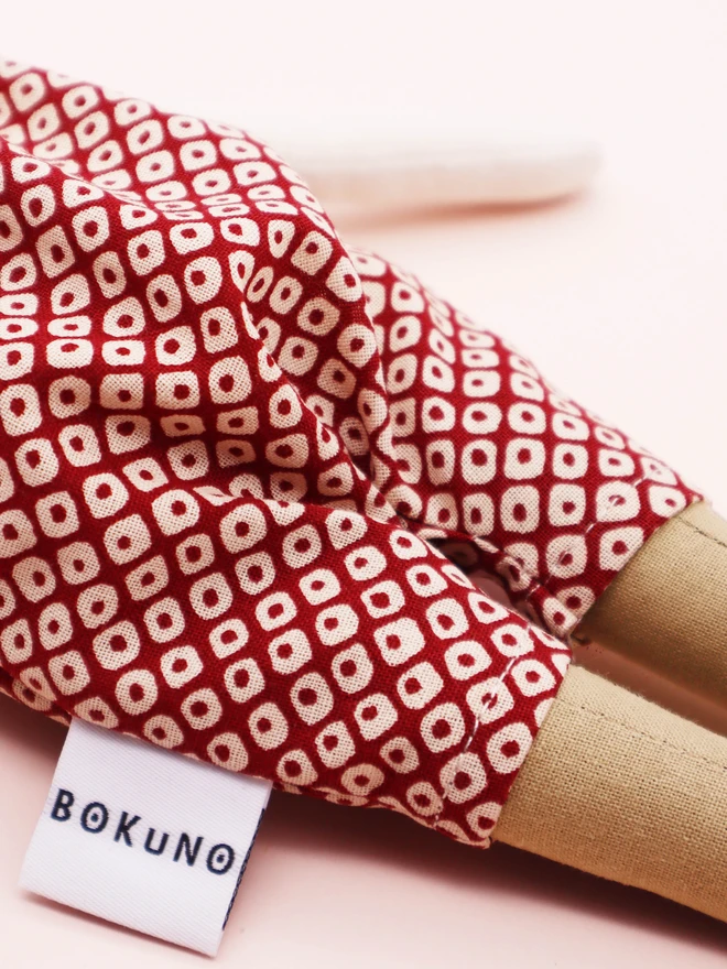 A handmade cloth doll with a light skin face, simple embroidered facial features, and black fabric hair styled with a matching patterned bow. The doll wears a red romper with a Japanese red shibori dots pattern.