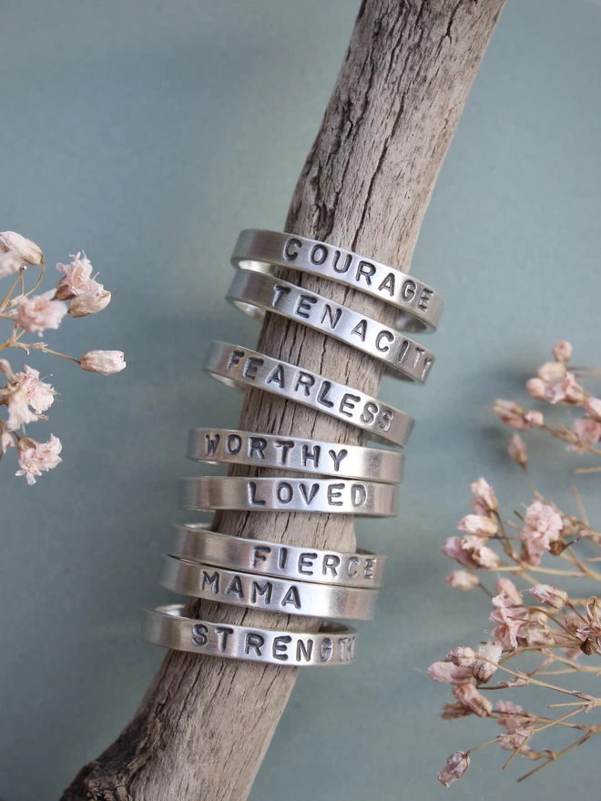 A selection of sterling silver rings stamped with empowering words, stacked on a driftwood stick held above a green background with tiny pink flowers on either side.