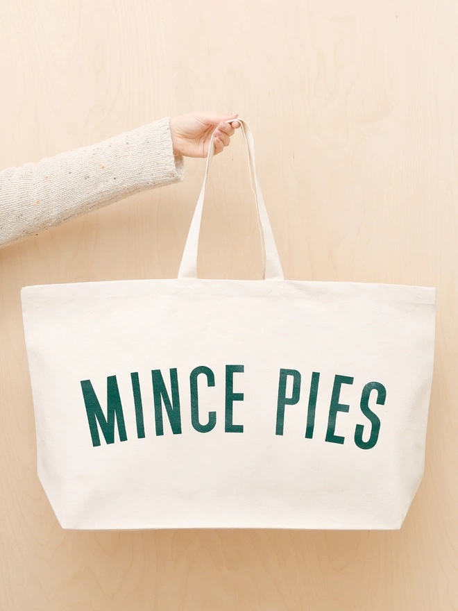 a hand holding out an oversized natural canvas tote bag with bold mince pies slogan