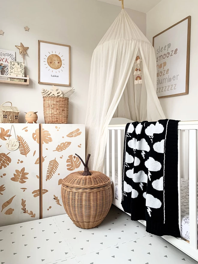 A neutral scandi style nursery with lots of natural wood, wicker accessories and a white cot. Over the cot is draped a black and white storm cloud baby blanket showing the reverse colourway.