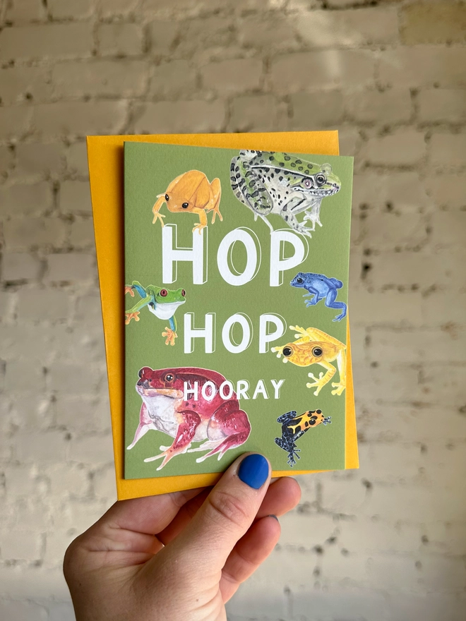 a green greetings card featuring a selection of frog illustrations with the text “hop hop hooray” with a deep yellow envelope