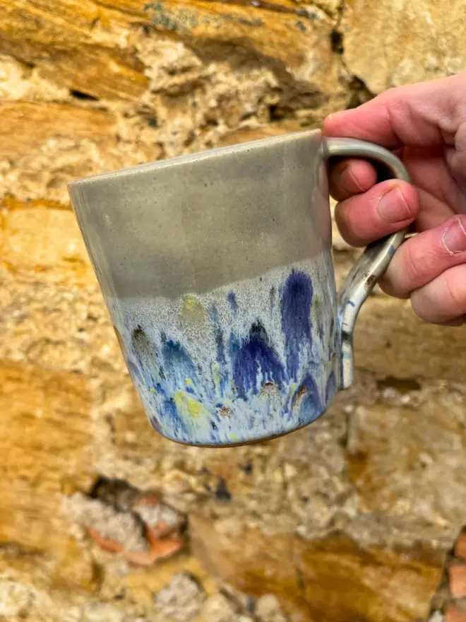 Shoreline Standard Mug, a colourful mug held in front of a brick wall. 
