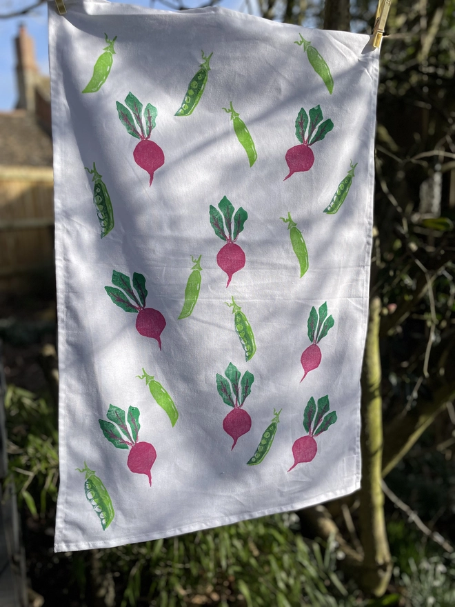Beetroot And Pea Kitchen Tea Towel. White fabric with a diagonal pattern of green pea pods and beetroots, some open to show bright green peas.