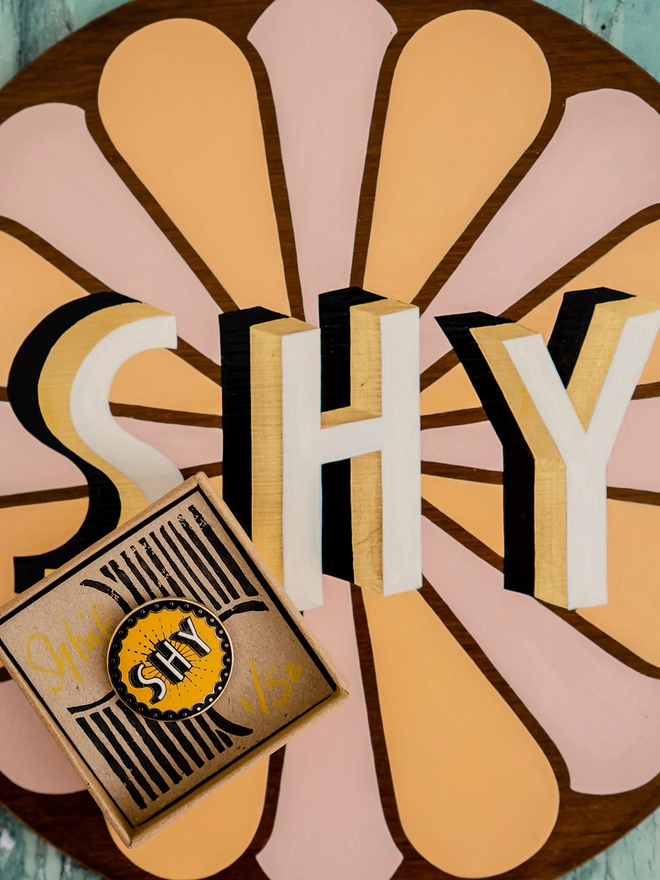 SHY cloisonne brooch in a hand printed gift box, on top of a painting of the word shy.