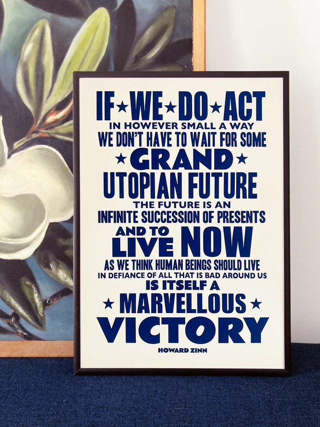  Framed typographic print, with navy blue text on a white background. Quote by Howard Zinn - "If we do act, in however small a way, we don't have to wait for some grand Utopian future, the future is an infinite succession of presents, and to live now as we think human beings should live, in defiance of all that is bad around us, is itself a marvellous victory." The print rests against a painting of white flowers on a dark blue background.