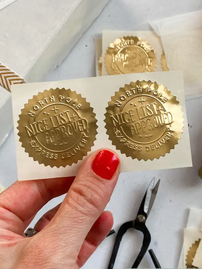 A hand holding a sheet of embossed gold foil seal stickers reading "North Pole, Nice List Approved, Express Delivery", with more "Lapland Approved" seals behind.