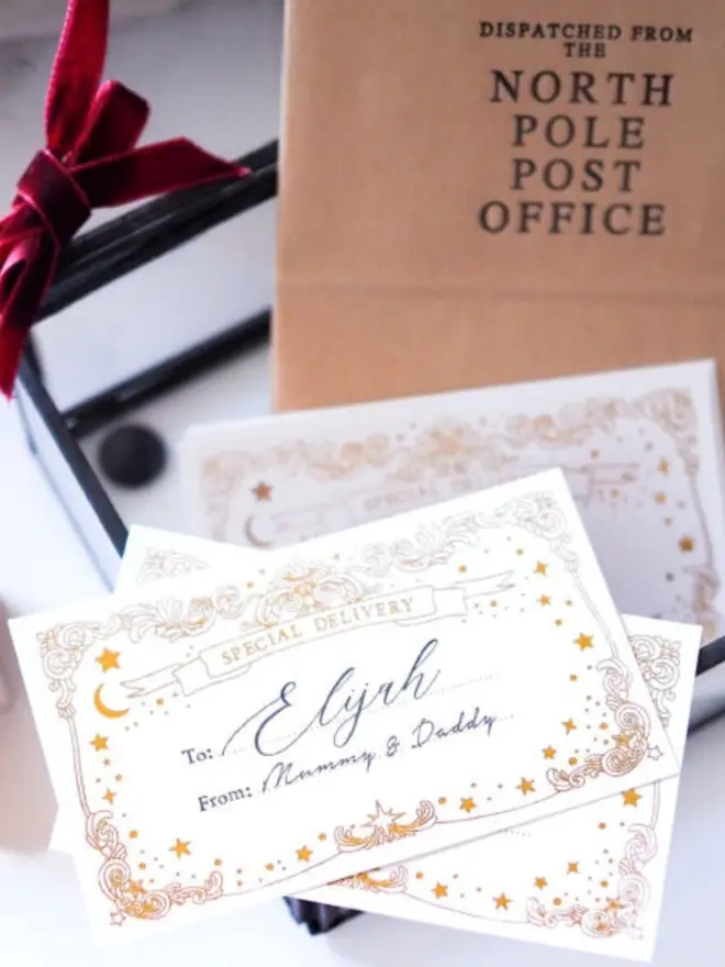 "Special Delivery" Christmas notelets with gold foil borders of scrolls, stars and a crescent moon, one handwritten "To: Elijah, From: Mummy & Daddy". Behind them are a gift tied with a burgundy velvet ribbon and a kraft paper bag printed "Dispatched from the North Pole Post Office".