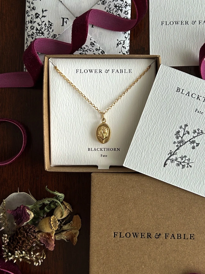 Gold blackthorn medal charm on a belcher chain necklace with flower & fable packaging on a darkground