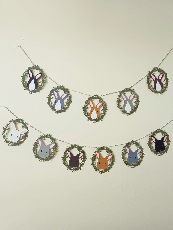 Easter Bunny Garland (5 bunny option and 6 bunny option) hung on plain cream wall.  5 bunny garland features a mix of brown, black and grey rabbit faces with white noses and pink ears. 6 Bunny garland features brown black grey and white rabbit faces with pink ears 