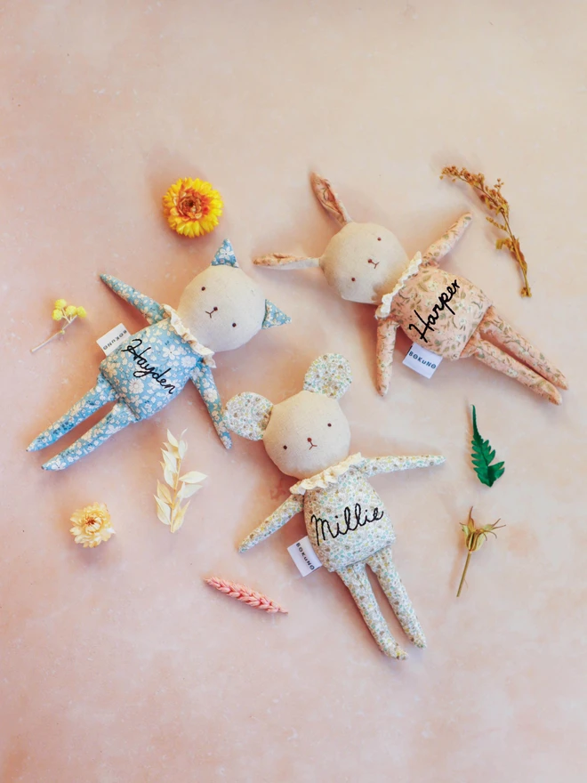 Three handmade fabric dolls with pastel floral prints: a cat, a mouse, and a rabbit. Each has a linen face, embroidered features, ruffled collars, and a 'BOKUNO' tag, highlighting their artisan craftsmanship. 