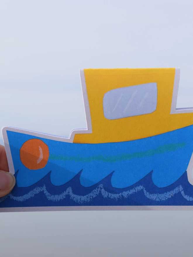 A cut out card of a boat with the sea in the background