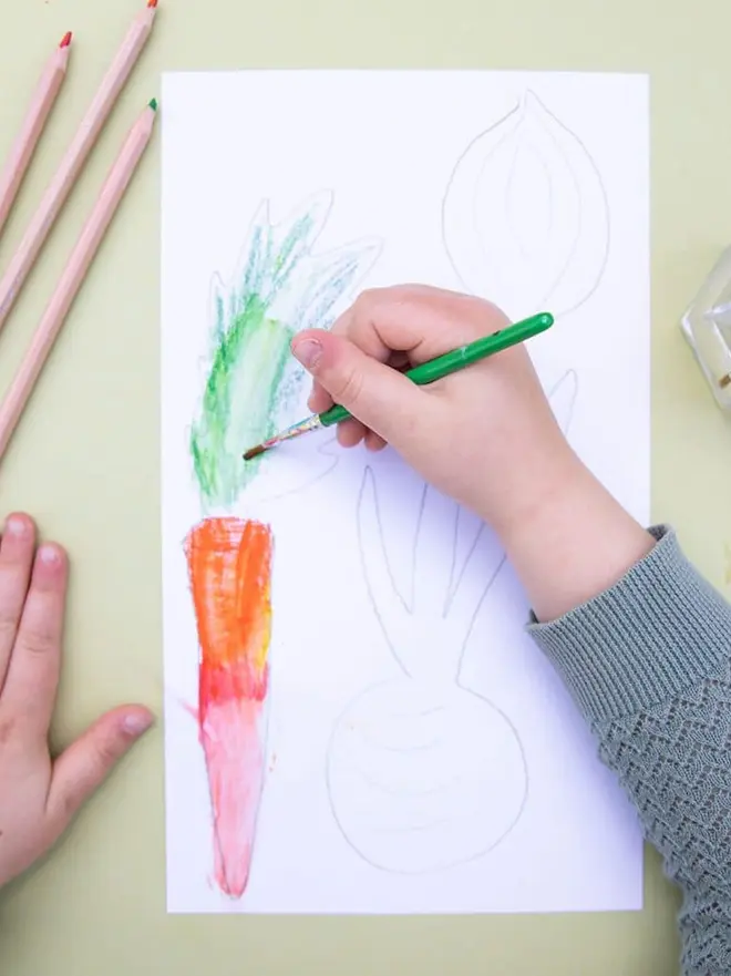in the garden mini activity box for children. a child's hand drawing a carrot and other veg on paper.