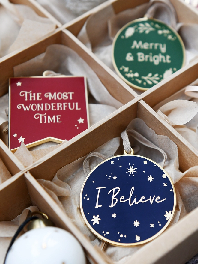 Three enamel Christmas decorations are tucked into a sectioned box. Each decoration has a traditional look with festive words on.