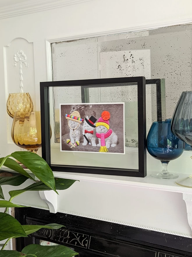 B&W print, 3 cats in embroidered sombrero, top hat and bobble hat, framed on mantlepiece