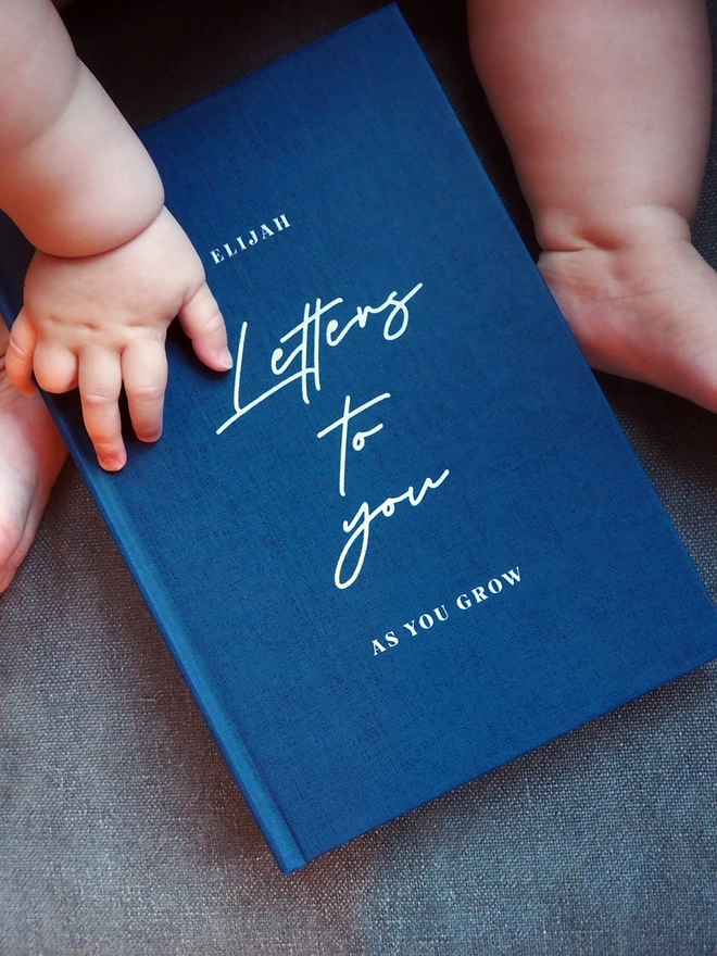 A navy journal being held by a baby with the words letters to you as your grow printed on the front
