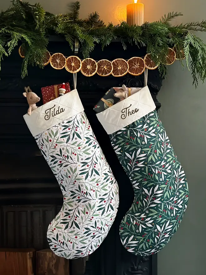 Two handmade quilted Christmas stockings hang on a fireplace decorated with greenery and orange slice garland.