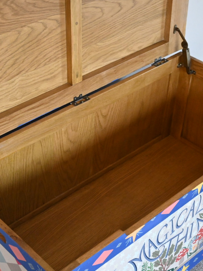 A slightly angled shot looking into an open wooden chest. The interior of the chest is a light, natural wood color with visible wood grain. The exterior of the chest is painted and features the words "Magical Things" in a whimsical font, along with colorful illustrations including mushrooms, flowers, and a diamond pattern. The hinges and support mechanism for the lid are visible. The chest is open, revealing its empty interior.