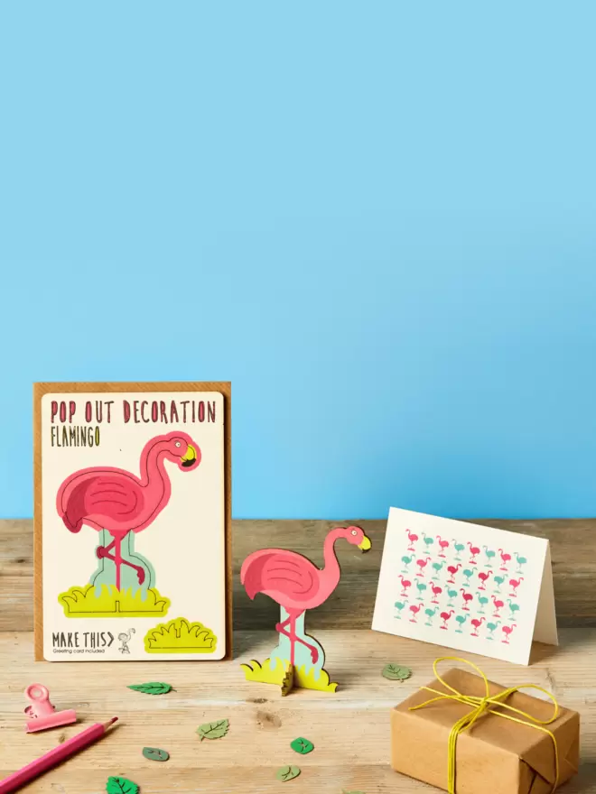3D laser-cut pink flamingo decoration and flamingo pattern greeting card and brown kraft envelope on top of a wooden desk in front of a sky blue coloured background