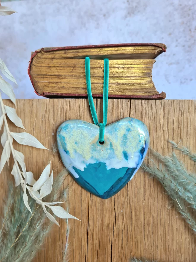 ceramic hanging heart decoration with blue, green, turquoise, Jenny Hopps Pottery, velvet ribbon, gift, mothers day gift, valentines gift, homeware, photographed hanging from an old book with dried flowers.