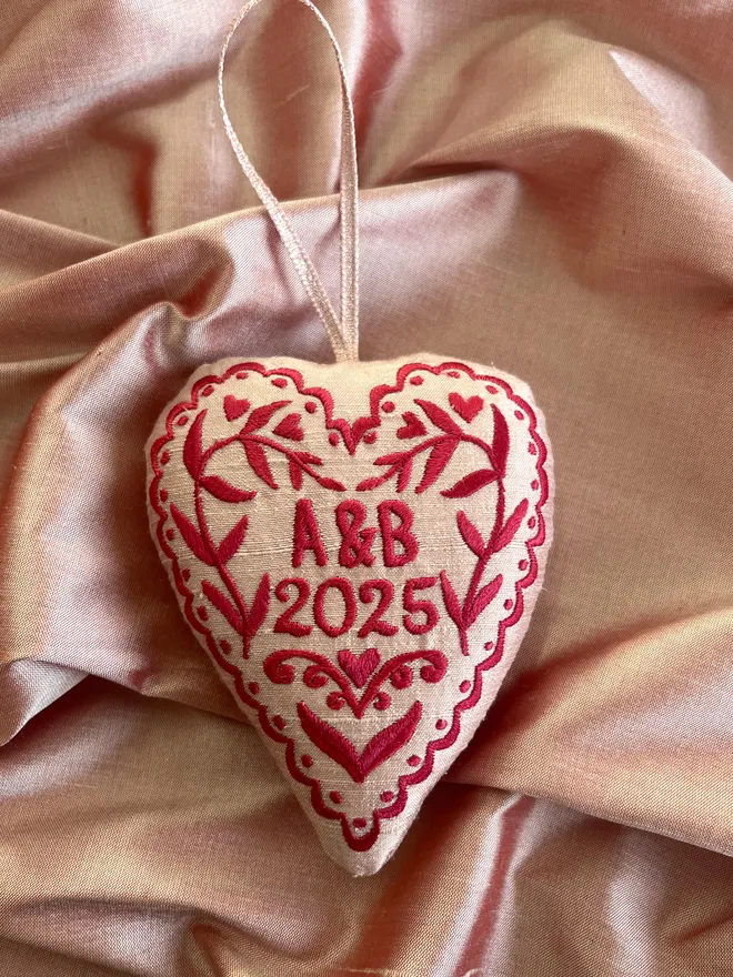 Pink silk stuffed heart ornament with a freehand embroidered design with initials and year in red thread with a pink metallic ribbon