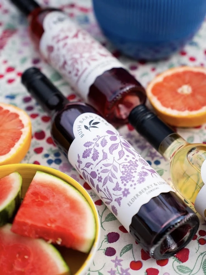 Elderberry & Lemon Cordial (Set Of 1, 2, 3 Or 4), a group of bottles of cordials sitting on a rug outside joined by some fruit. 