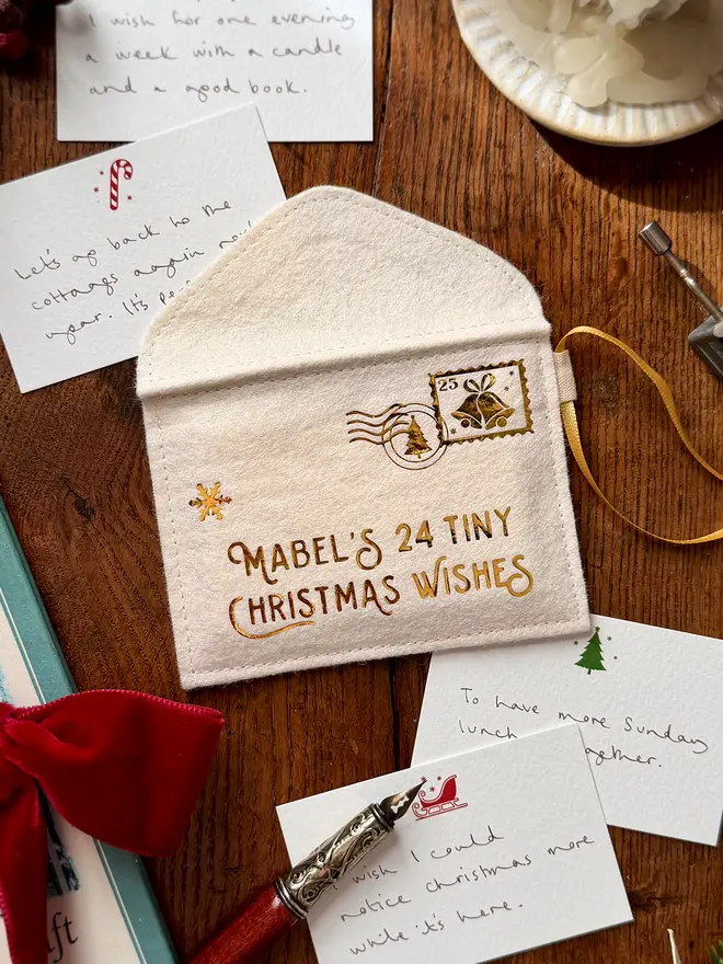 A handmade felt envelope advent calendar with golden lettering that reads 'Mabel's 24 Tiny Christmas Wishes' with several Christmas notecards all laying on a wooden table.