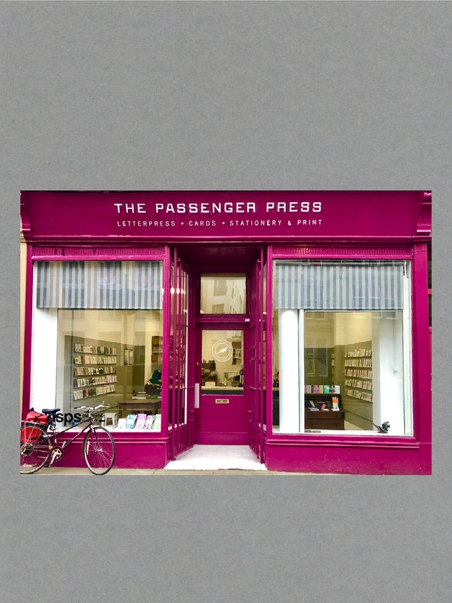 Raspberry pink shop front with a sign reading ‘The Passenger Press’ in white. A bicycle leans against the shop window in the street in front.