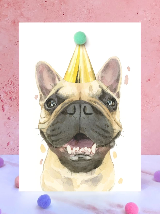 A greeting card featuring a hand painted design of a fawn coloured French bulldog, stood upright on a marble surface surrounded by pompoms. 