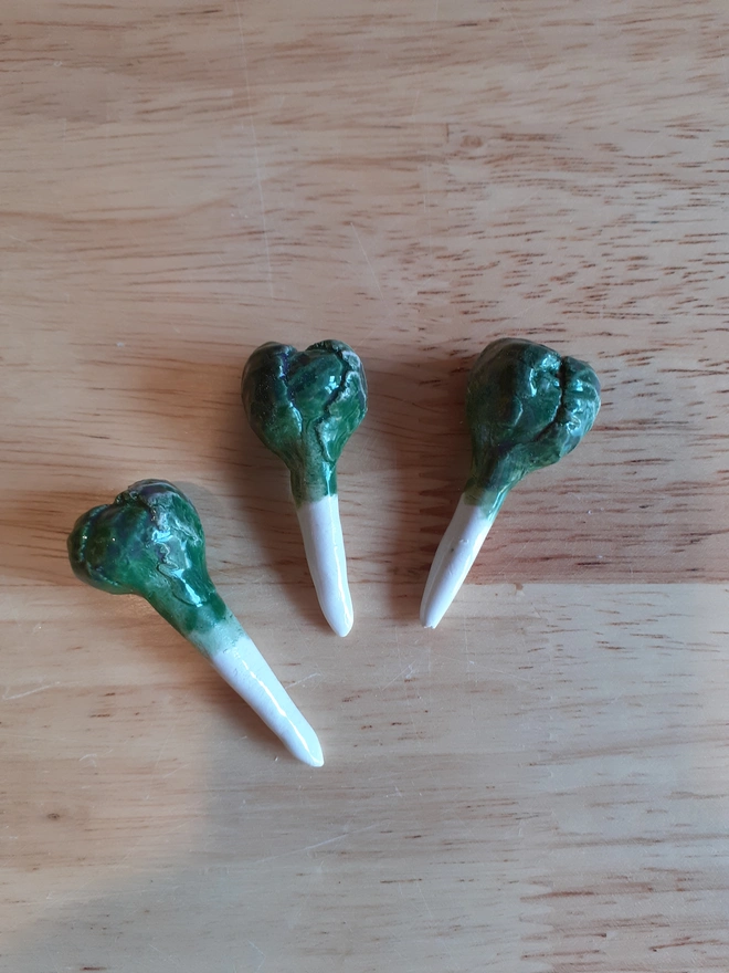 Set of three handmade ceramic Brussels sprout candle holders laid on a wooden surface, showing their leafy green glaze and pointed white stems.