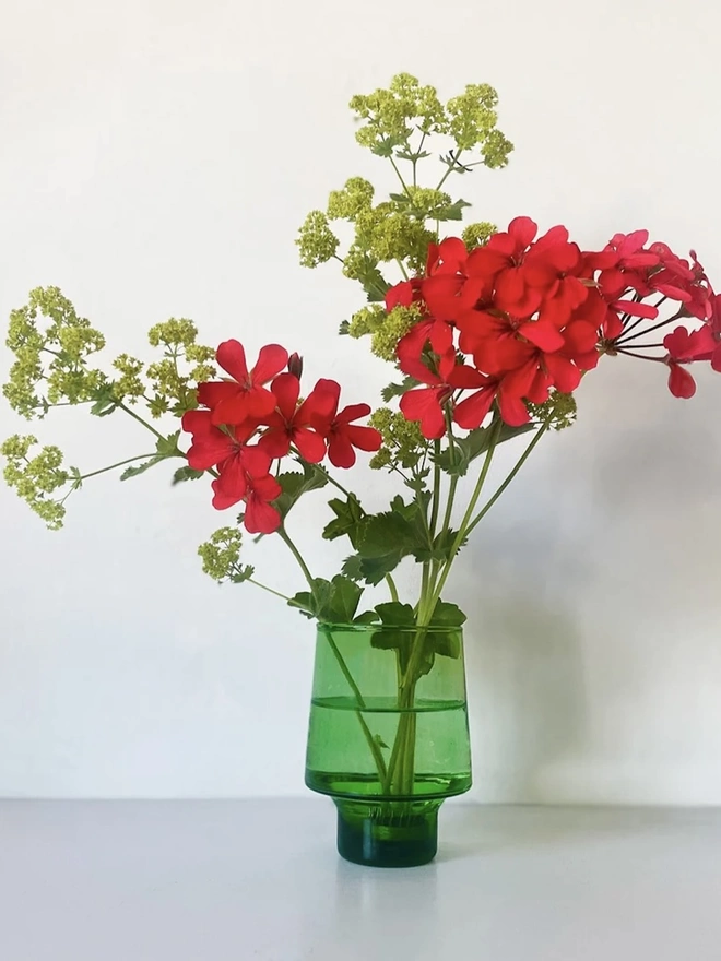 Tall Green Flower Arranging Items (Set Of 2), a green vase sitting on a grey surface against a white backdrop. It is filled by a bundle of red flowers held together by a copper frog. 