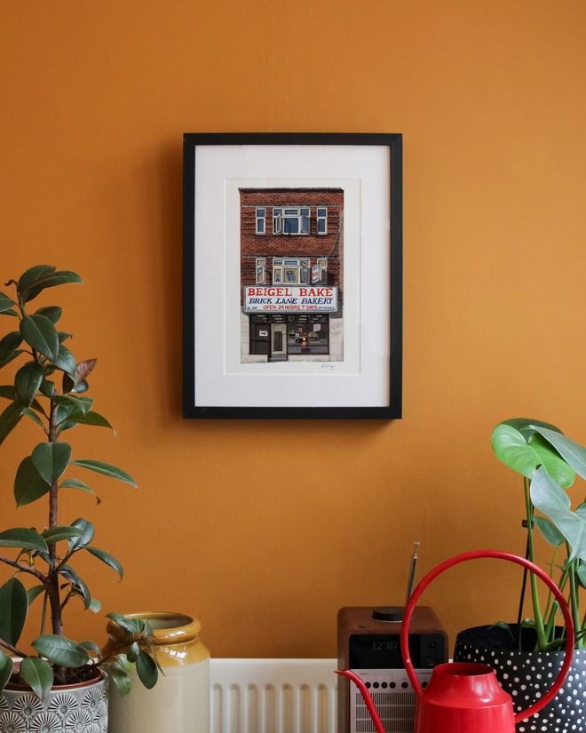 An illustration of Beigel Bake in a black frame, centered on a warm orange wall above indoor plants, a vintage radio, and a red watering can.
