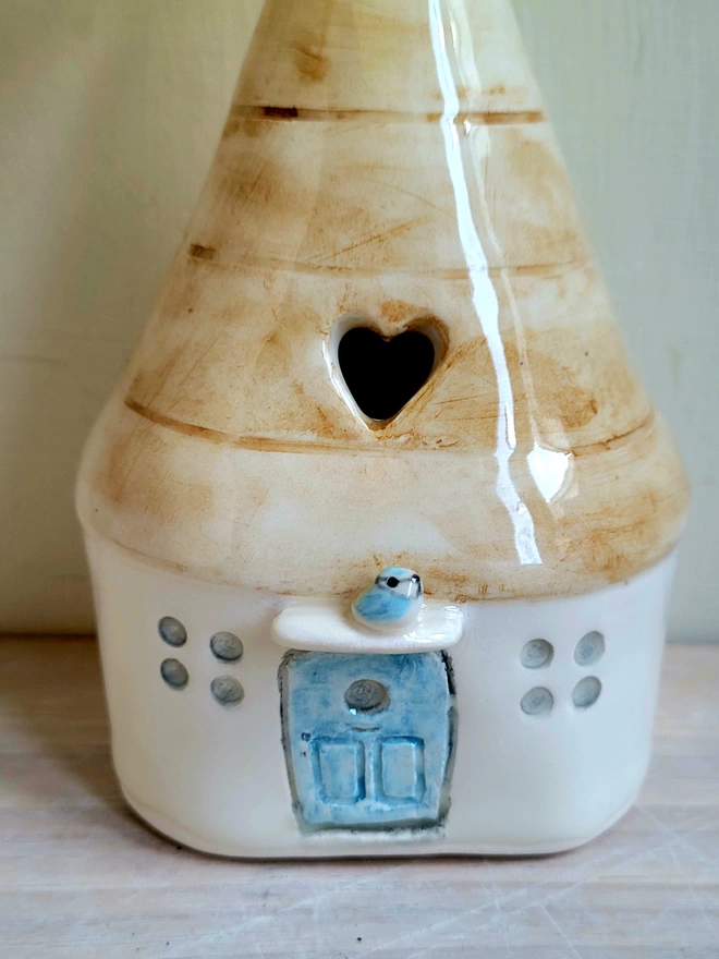 view of a ceramic house vase on a wooden counter with a blue door with a modelled blue tit above and a heart cut out of the brown roof