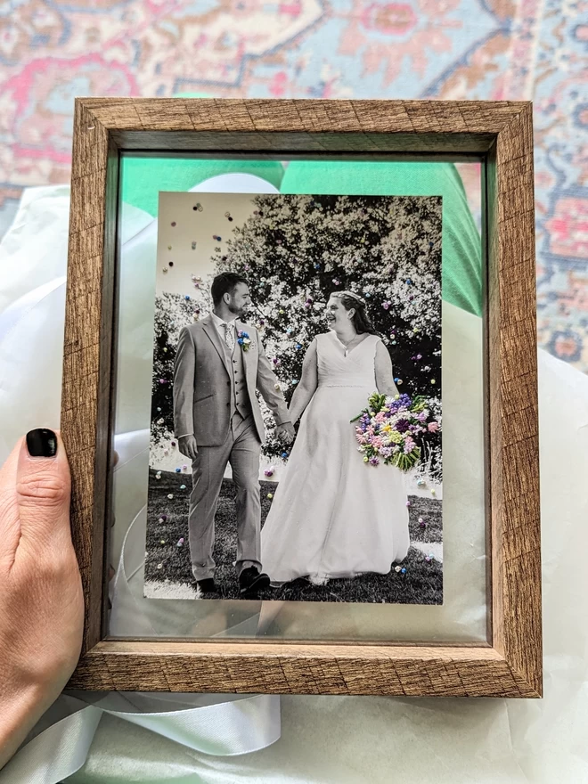 Embroidered floral bouquet and confetti on photo held in wooden double glass frame 