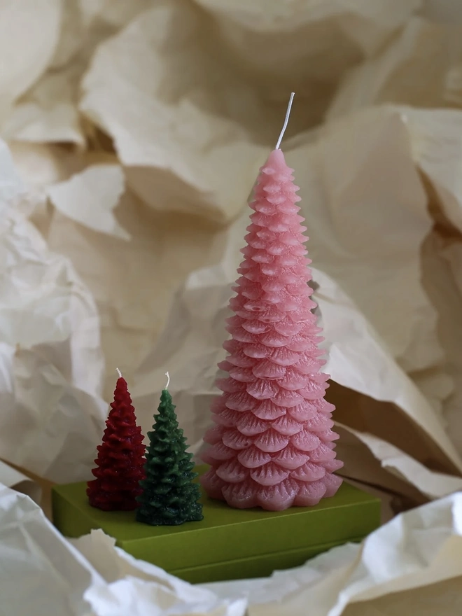 Large Christmas Tree Candle. a large pink christmas tree shaped candle stands on a green box, nestled among crumpled cream tissue paper. a smaller red and a smaller dark green tree candle sit in front of it for scale, both with visible wicks. the textured, layered pine branch design and slightly variegated wax give all three candles a realistic, hand-poured finish.