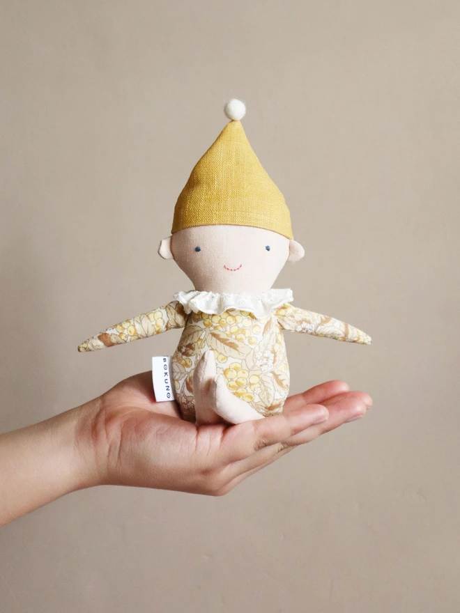 A handmade Spring Elf Doll with fair skin, a yellow floral print body, and a mustard-yellow hat with a white pompom. The doll has a sweet smile and a delicate white ruffled collar.