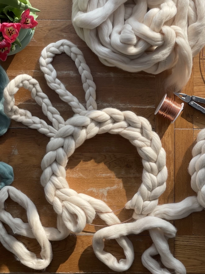 Taken from above, an oyster cream woolly rabbit wreath is being made on an oak table, the sunlight casting across the table and making the copper wire used for the bunny ears glint. 