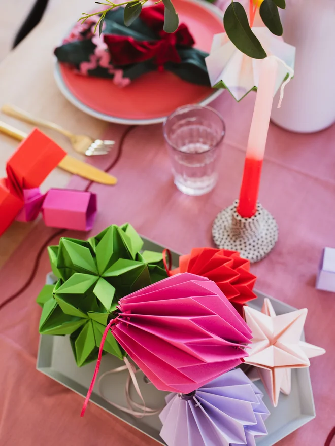 A Christmas table decorated with bright colourful paper shapes, a candle and place settings ready.