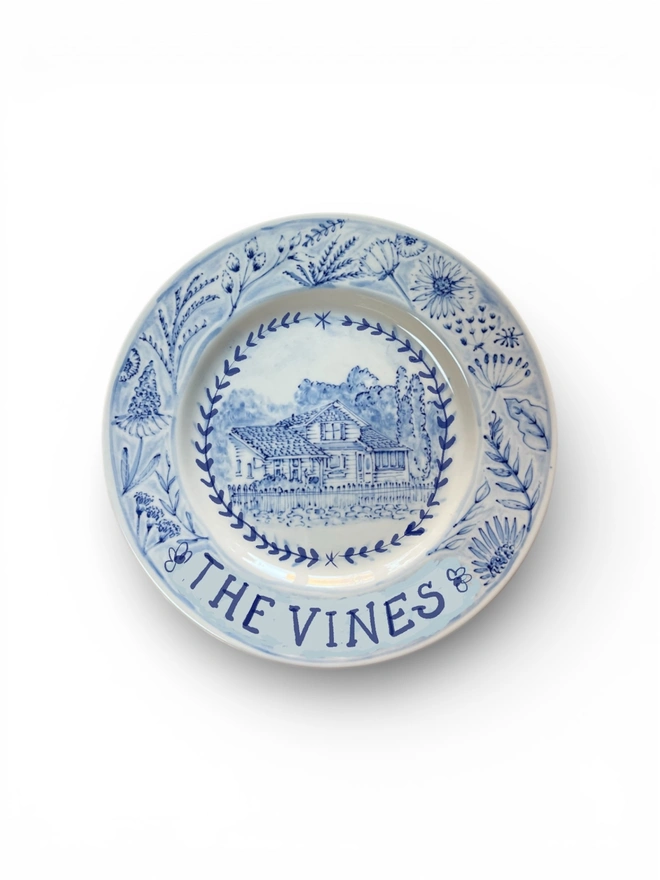 Blue and White hand painted plate with a portrait of a large house, with name "The Vines" underneath and a botanical border in a watercolour and illustration style