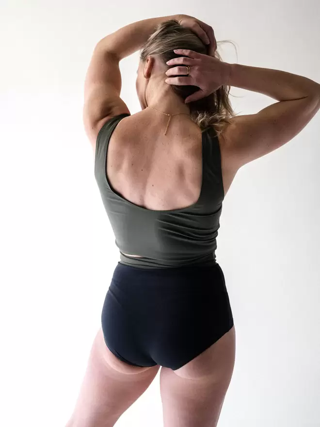 A back view of a blonde lady with her hands running through her hair, wearing a Davy J Sustainable Waterwear olive cropped swim top with a scoop back. She also wears a pair of black high waist bikini briefs with a rolled down waist to show olive lining