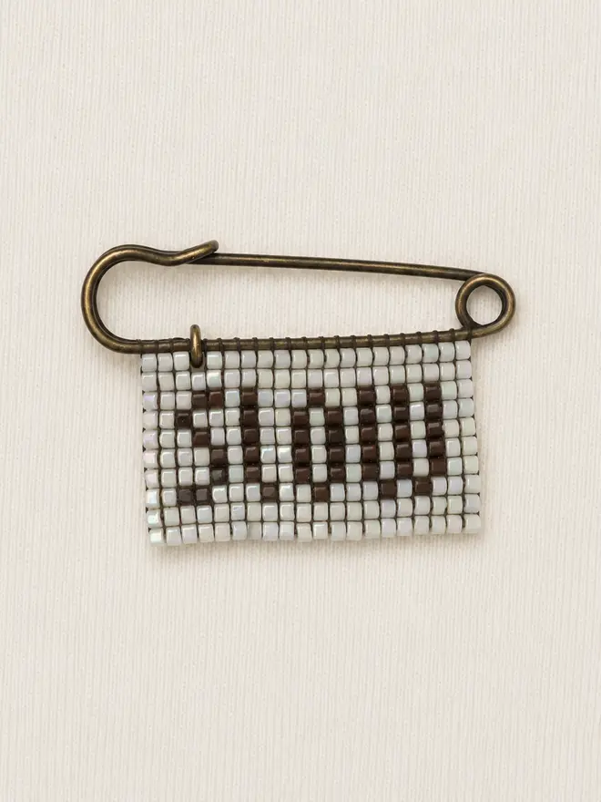 beaded 'slow' pin. a beaded pin badge with the word slow in brown lettering on a pearl background.