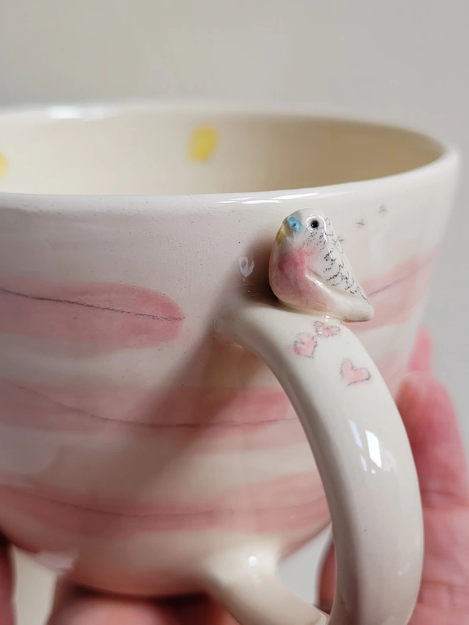 close up detail of budgie with pale pink chest on a  stripey pink and white pottery cup with pink hearts on the handle