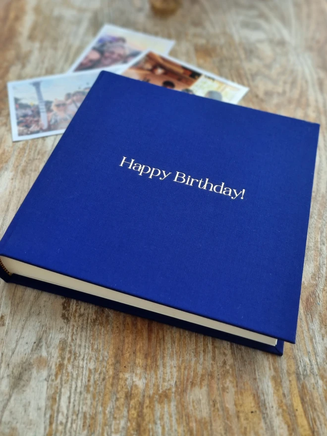 An indigo blue photo album printed with 'Happy Birthday!' in gold lettering sits on a wooden tabletop. There are photos in the background.